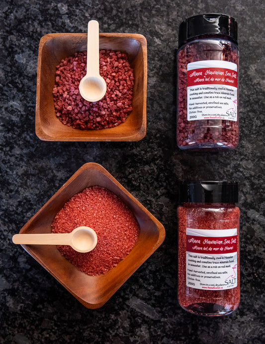 Alaea Hawaiian Sea Salt by The Salt Cellar displayed in two wooden bowls and matching spice jars. One bowl contains coarse, chunky salt crystals, while the other holds finer granules, both infused with rich red Alaea clay. This unrefined, hand-harvested sea salt is packed with trace minerals and is perfect for seasoning, meat rubs, and Hawaiian-inspired dishes. Gluten-free and free from additives or preservatives