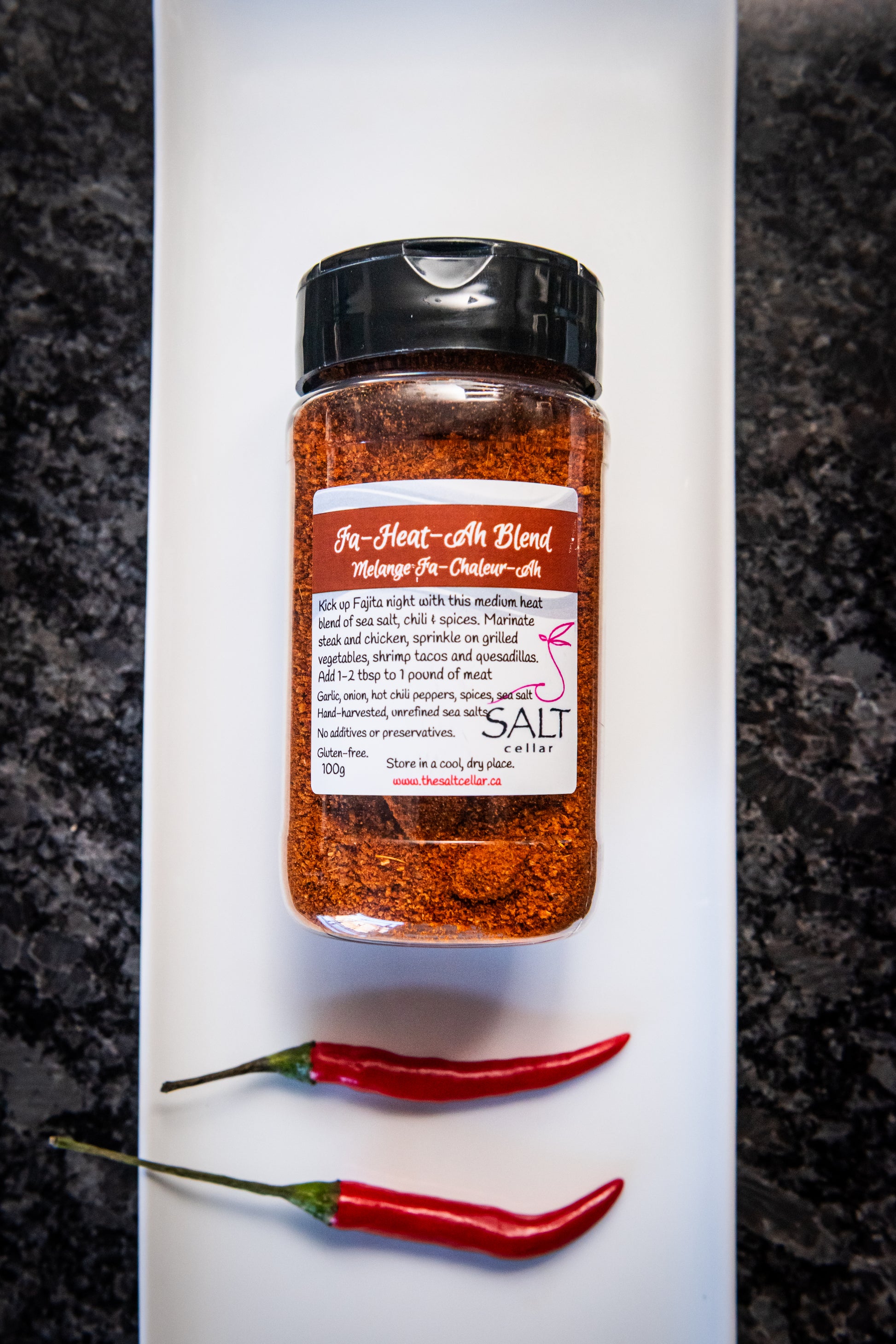 Fa-Heat-Ah Blend seasoning by The Salt Cellar displayed on a white plate with two fresh red chili peppers. This medium-heat spice blend, made with sea salt, chili, garlic, onion, and spices, is perfect for fajitas, steak, chicken, grilled vegetables, and shrimp tacos. Gluten-free, no additives, and handcrafted with unrefined sea salt.