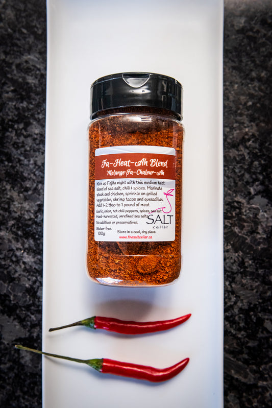 Fa-Heat-Ah Blend seasoning by The Salt Cellar displayed on a white plate with two fresh red chili peppers. This medium-heat spice blend, made with sea salt, chili, garlic, onion, and spices, is perfect for fajitas, steak, chicken, grilled vegetables, and shrimp tacos. Gluten-free, no additives, and handcrafted with unrefined sea salt.