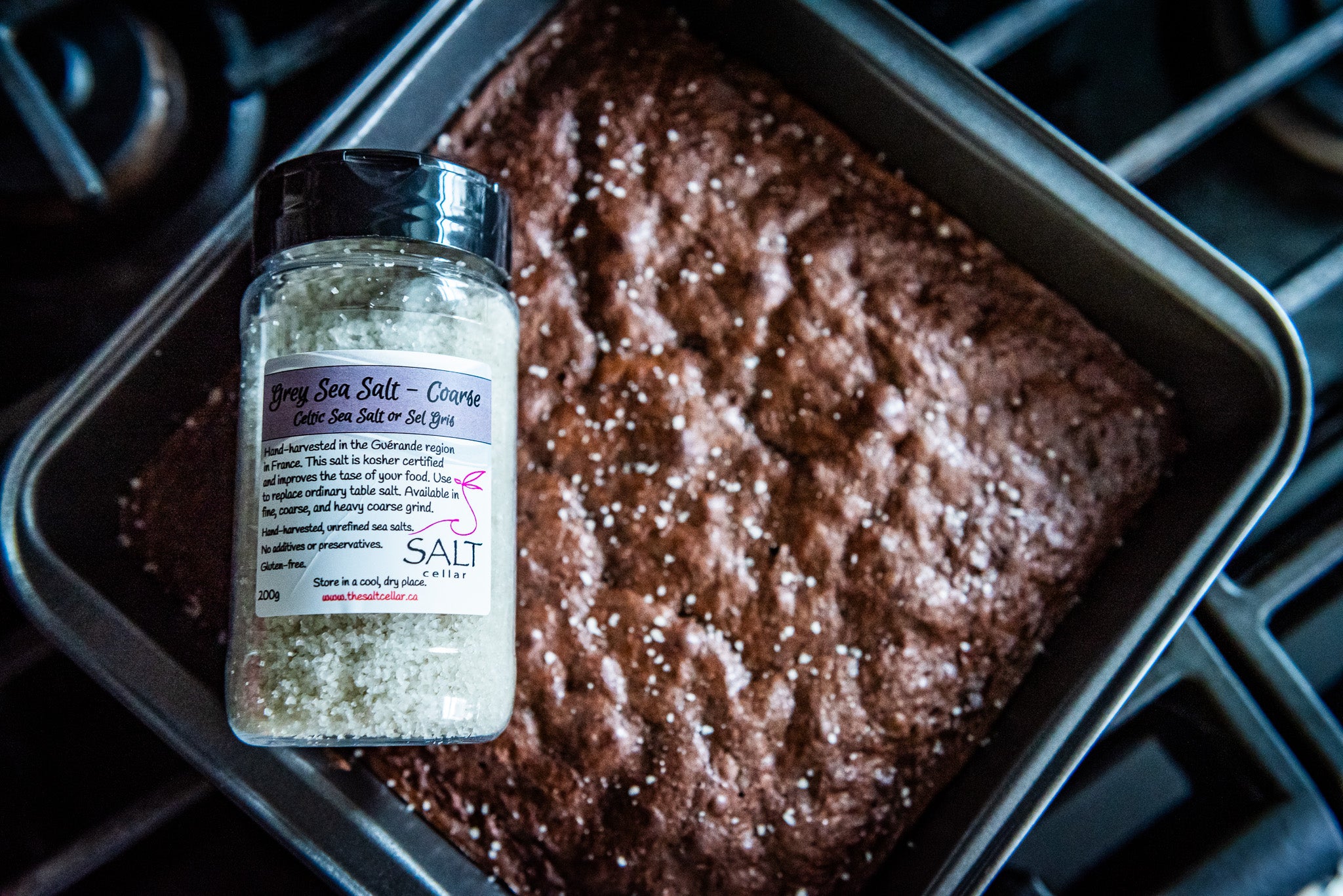 A fresh batch of homemade brownies sprinkled with The Salt Cellar’s Grey Sea Salt – Coarse sits in a baking pan, fresh from the oven. The jar of hand-harvested Celtic sea salt, sourced from the Guérande region of France, is prominently displayed in the foreground, highlighting its rich mineral content and flaky texture. The contrast of sweet chocolate and savory sea salt enhances the depth of flavor, making for a gourmet dessert experience.