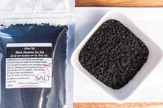 Hiwa Kai Black Hawaiian Sea Salt by The Salt Cellar, featuring jet-black, hand-harvested Pacific sea salt infused with activated charcoal from coconut shells. The salt is displayed in a resealable 200g bag and a white dish showcasing its coarse, irregularly shaped granules. Perfect for use in a grinder, as a finishing salt for seafood and meats, or as a bold cocktail rim garnish. Naturally rich in minerals, gluten-free, and free from additives or preservatives.