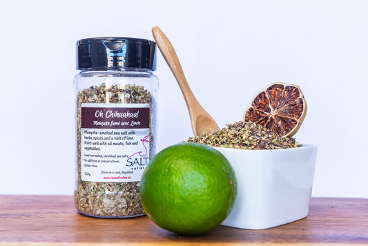 A close-up of The Salt Cellar's "Oh Chihuahua!" seasoning blend, featuring mesquite-smoked sea salt with herbs, spices, and a hint of lime. The spice blend is displayed in a white bowl with a dried lime slice, a wooden spoon, and a fresh lime, highlighting its zesty, smoky flavor. The seasoning jar is placed beside it, showcasing the label's details.