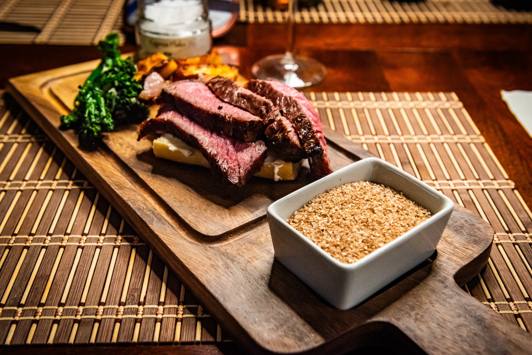 A beautifully plated steak with slices of perfectly cooked, medium-rare beef sits on a wooden serving board. A side of crispy broccolini and roasted potatoes completes the dish. In the foreground, a small dish of golden smoked sea salt from The Salt Cellar adds a gourmet touch, enhancing the rich flavors of the meal. A jar of seasoning and a glass of wine in the background set the scene for a refined dining experience.