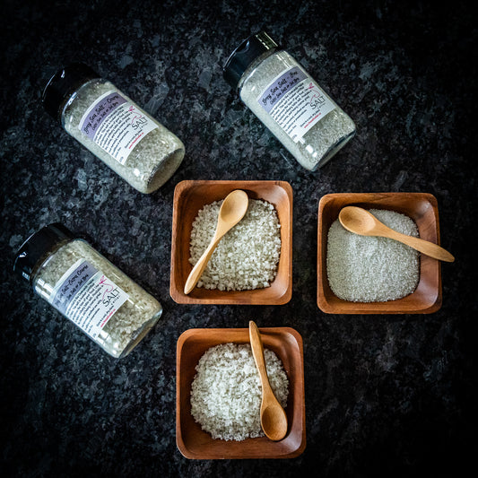 A selection of The Salt Cellar’s all-natural grey sea salts, also known as Celtic sea salt, are displayed on a dark granite surface. Three wooden bowls contain different textures of grey sea salt, each accompanied by a small wooden spoon. Surrounding the bowls are jars of Grey Sea Salt, showcasing their coarse, mineral-rich texture. These gourmet salts are perfect for enhancing the flavor of meats, vegetables, and other dishes.